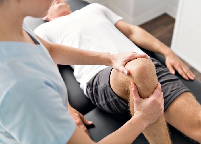 RUNWAY HEALTH - Knee Pain Exam patient in white t-shirt on physio table laying face up with a physiotherapist examinin right knee pain with knee flexed.
