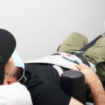 Man with a black hat, shirt and green pants laying backside down on a table receiving spinal decompression therapy