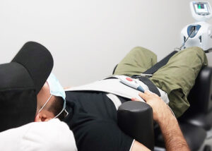 Man with a black hat, black shirt and green pants laying backside down on a table receiving spinal decompression therapy. He seems to be quite comfortable.