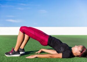 Dark haired woman with pink yoga and black shirt, laying down on green turf doing a glute-bridge exercise