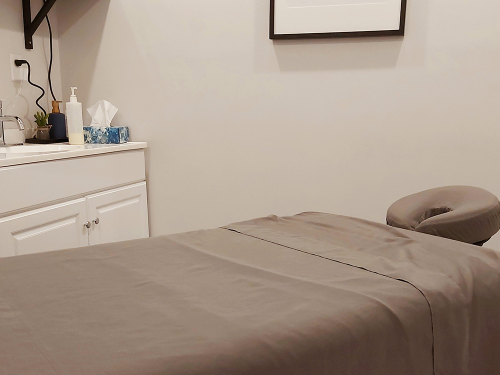 Flat massage therapy bed with grey sheets in a room with white walls.