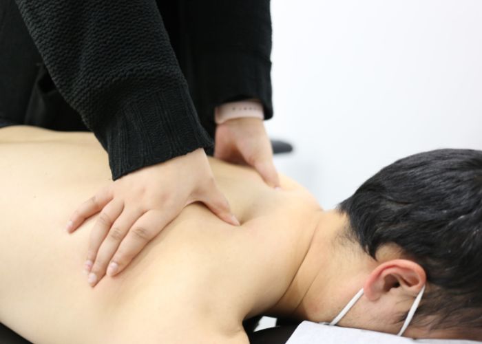 Dr. Marina, a chiropractor is applying pressure to the upper back muscles of a male patient who is lying on their stomach on a chiropractic table.