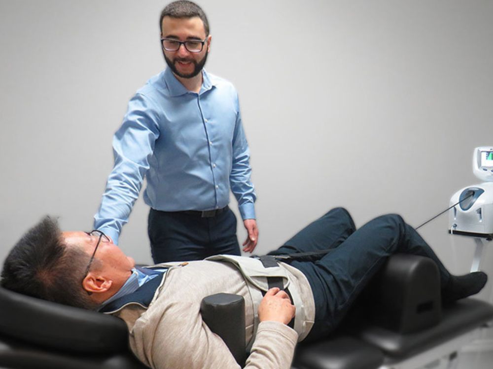 A middle aged man is receiving spinal decompression therapy at Runway Health, a clinic located either in Markham or Newmarket. The patient is being comforted by a chiropractor Dr. Omar. Spinal decompression is a great treatment technique to help alleviate pain from lumbar osteoarthritis, disc herniations, disc bulges, sciatica, and spinal stenosis.